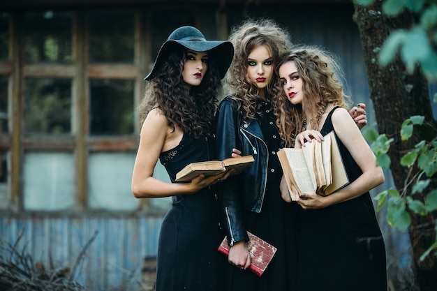 Tres mujeres vintage como brujas, posan frente a un edificio abandonado con libros en la mano en la víspera de Halloween
