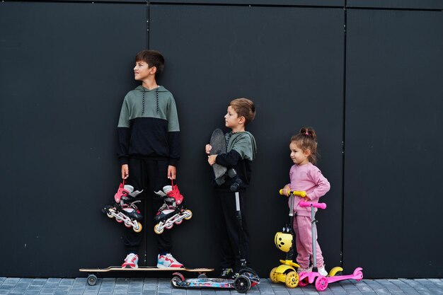 Tres niños al aire libre contra la pared moderna negra La familia deportiva pasa tiempo libre al aire libre con scooters y patines