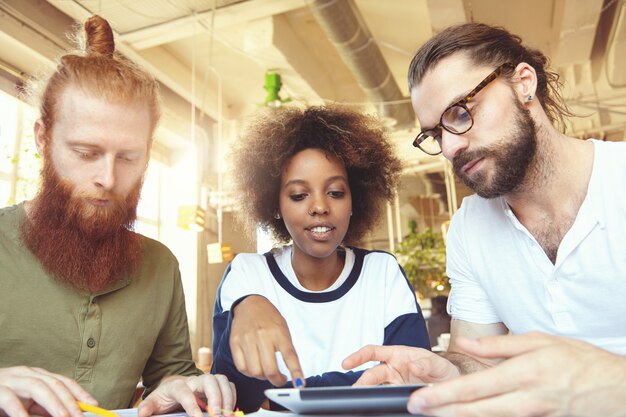 Tres personas creativas que tienen una discusión en el café: mujer africana explicando su visión, señalando la pantalla del panel táctil, hombre barbudo con gafas escuchando atentamente y pareja pelirroja tomando notas