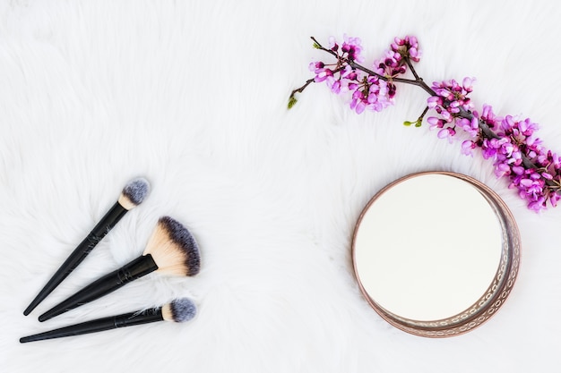 Tres pinceles de maquillaje con espejo redondo y ramita de flor artificial sobre fondo de piel blanca