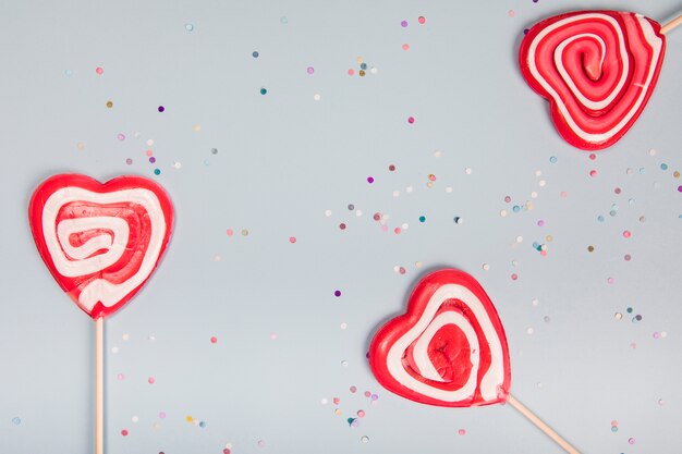 Tres piruletas rojas de la forma del corazón en fondo gris con confeti colorido