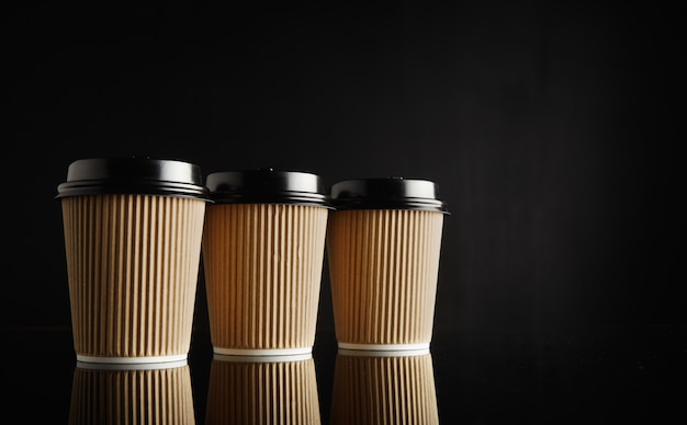 Tres tazas de café para llevar de cartón marrón claro idénticas con tapas negras en una fila en la mesa negra reflectante contra la pared negra