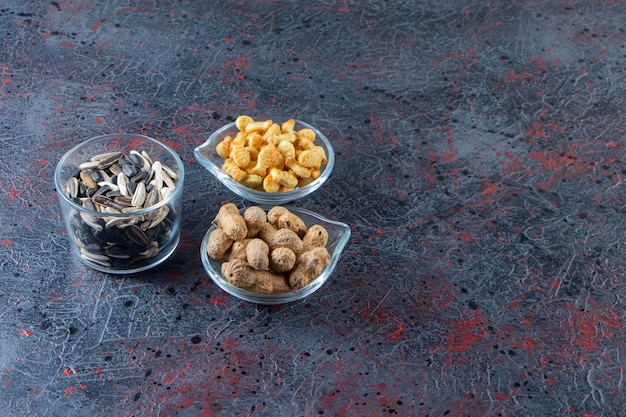 Tres tazones de cacahuetes, semillas de girasol y galletas saladas sobre fondo azul.