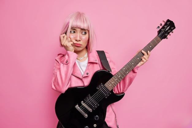 Triste y aburrida rockera sostiene una guitarra acústica eléctrica crea una nueva canción para su nuevo álbum tiene un peinado rosa vestido con una chaqueta