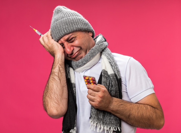 Foto gratuito triste adulto enfermo hombre caucásico con bufanda alrededor del cuello con gorro de invierno sosteniendo jeringa y medicina blister aislado en la pared rosa con espacio de copia