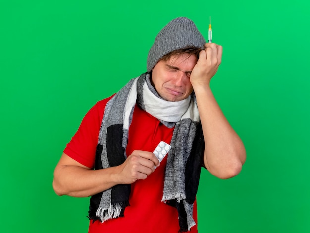 Foto gratuita triste y asustado joven guapo rubio enfermo con gorro de invierno y bufanda sosteniendo un paquete de tabletas médicas y una jeringa tocando la cabeza con los ojos cerrados aislados sobre fondo verde con espacio de copia