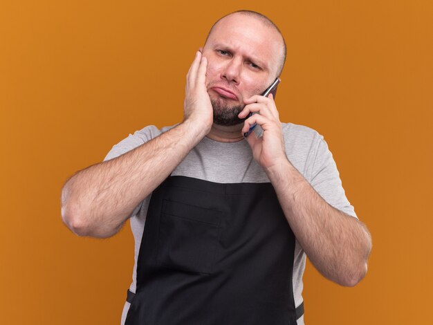 Triste barbero masculino de mediana edad eslavo en uniforme habla por teléfono poniendo la mano en la mejilla aislada en la pared naranja