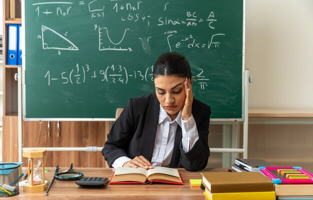 Triste con la cabeza baja joven maestra se sienta a la mesa con útiles escolares poniendo la mano en el templo en el aula