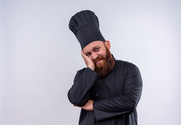 Un triste chef barbudo en uniforme negro pensando y manteniendo la mano en la cara en una pared blanca