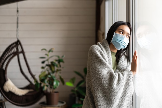 Triste chica asiática en mascarilla médica mirando fuera de la ventana y anhelo ir a caminar estando enferma catchi ...