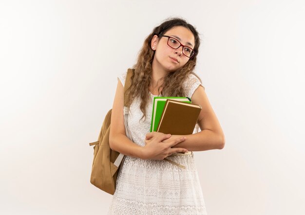 Triste colegiala bonita joven con gafas y mochila sosteniendo libros mirando al lado aislado en la pared blanca