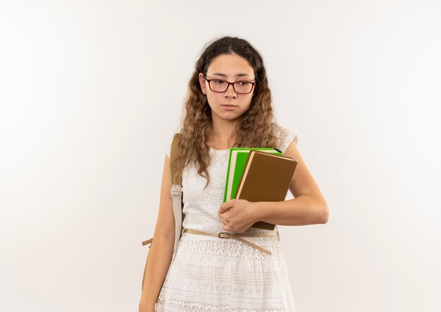 Triste colegiala bonita joven con gafas y mochila sosteniendo libros mirando al lado aislado en la pared blanca