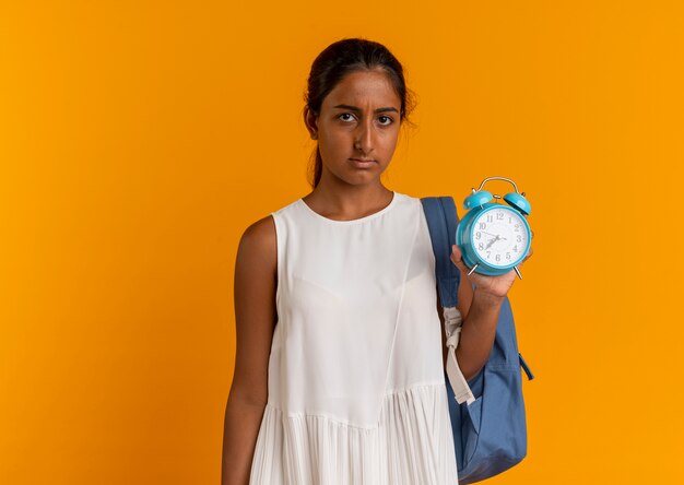 Triste colegiala joven vistiendo bolsa trasera con reloj despertador en naranja
