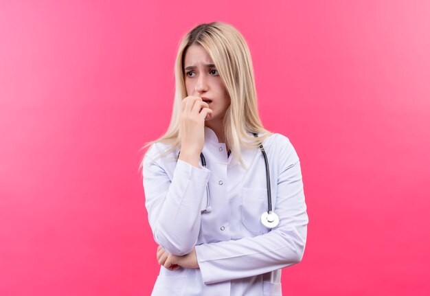 Triste doctor joven rubia con estetoscopio en bata médica puso su mano en la barbilla sobre fondo rosa aislado