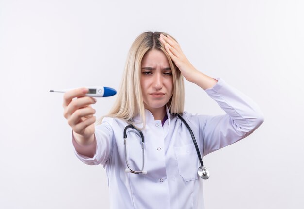 Triste doctor joven rubia con estetoscopio y bata médica sosteniendo el termómetro a la cámara puso su mano una cabeza sobre fondo blanco aislado