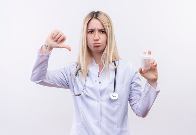 Triste doctor joven rubia con estetoscopio y bata médica sosteniendo vacía puede su pulgar hacia abajo sobre fondo blanco aislado