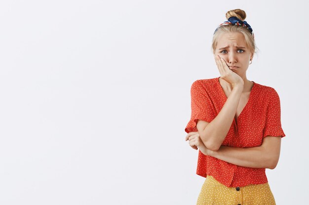 Triste e incómoda joven rubia posando contra la pared blanca