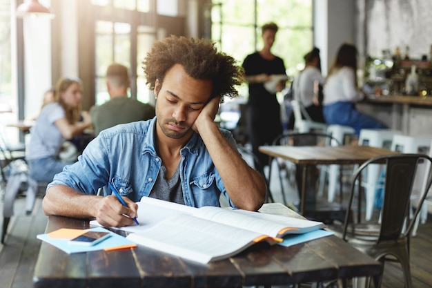 Triste e infeliz estudiante barbudo de piel oscura que se siente frustrado mientras se prepara para las lecciones en la universidad, escribe en su cuaderno con un bolígrafo, se apoya en el codo y mira notas con expresión molesta