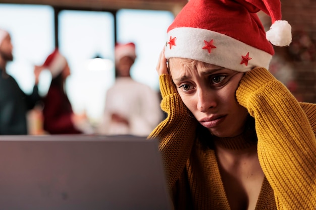 Foto gratuita triste e infeliz mujer caucásica con sombrero de papá noel trabajando en una computadora portátil en la oficina en vísperas de navidad. empleado de la empresa exhausto resolviendo tareas en la computadora durante la temporada de año nuevo