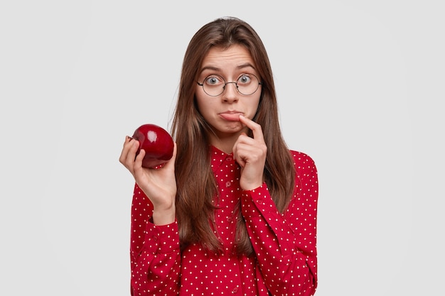 Triste y encantadora mujer vegetariana sostiene una manzana roja fresca, se frunce el labio inferior, mantiene una dieta saludable, come fruta, tiene el pelo largo y liso, vestida con una blusa de lunares