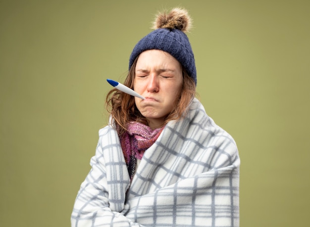 Triste enferma joven vestida con túnica blanca y gorro de invierno con bufanda envuelta en cuadros poniendo termómetro en la boca aislado en verde oliva