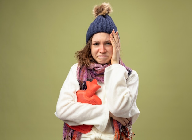 Triste enferma joven vestida con túnica blanca y gorro de invierno con bufanda sosteniendo una bolsa de agua caliente poniendo la mano en la cabeza aislada en verde oliva