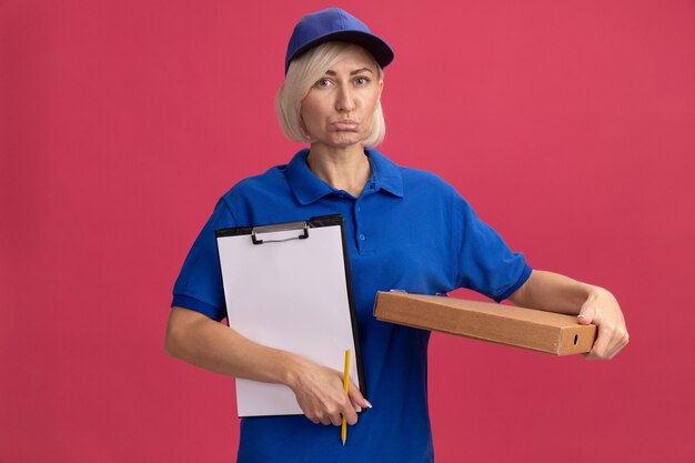 Triste entrega rubia de mediana edad en uniforme azul y gorra sosteniendo el paquete de pizza lápiz portapapeles aislado en la pared rosa con espacio de copia