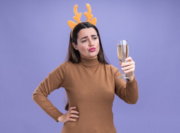 Triste hermosa joven vestida de suéter marrón con aro de pelo de Navidad sosteniendo y mirando una copa de champán poniendo la mano en la cadera aislada sobre fondo azul