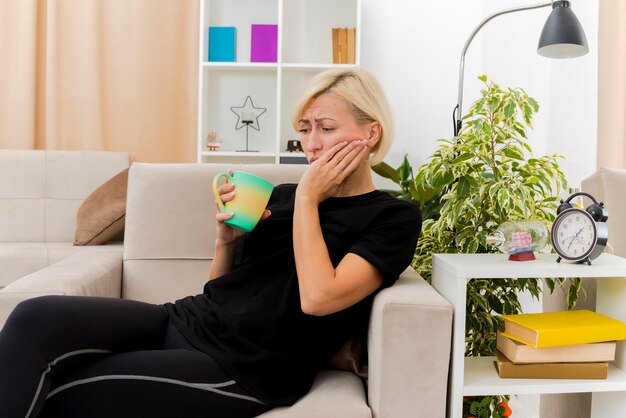 Triste hermosa mujer rusa rubia acostada en el sillón poniendo la mano en la cara sosteniendo la taza dentro de la sala de estar