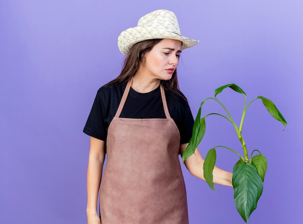 Triste hermosa niña de jardinero en uniforme con sombrero de jardinería sosteniendo y mirando la planta aislada en azul