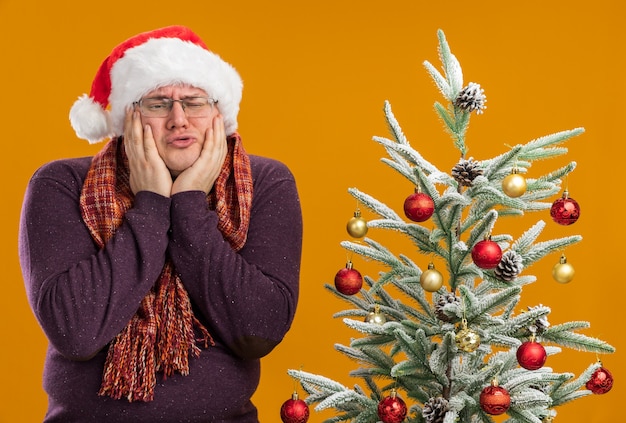 Triste hombre adulto con gafas y gorro de Papá Noel con bufanda alrededor del cuello de pie cerca del árbol de Navidad decorado manteniendo las manos en la cara mirando al lado aislado sobre fondo naranja