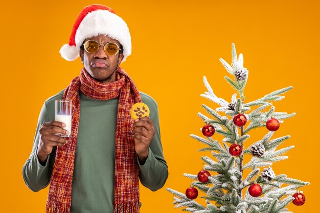 Triste hombre afroamericano en santa sombrero y bufanda alrededor del cuello sosteniendo un vaso de leche y galletas frunciendo los labios de pie junto a un árbol de Navidad sobre fondo naranja