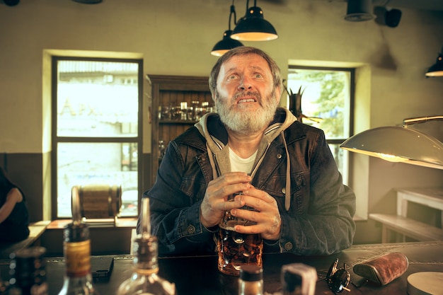 El triste hombre barbudo senior bebiendo cerveza en pub
