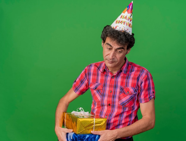 Triste hombre caucásico de mediana edad con gorra de cumpleaños sosteniendo paquetes de regalo mirando hacia abajo aislado sobre fondo verde con espacio de copia