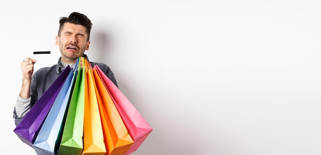 Foto gratuita triste hombre llorando desperdició todo el dinero en compras sosteniendo bolsas y mostrando una tarjeta de crédito vacía de pie de nuevo