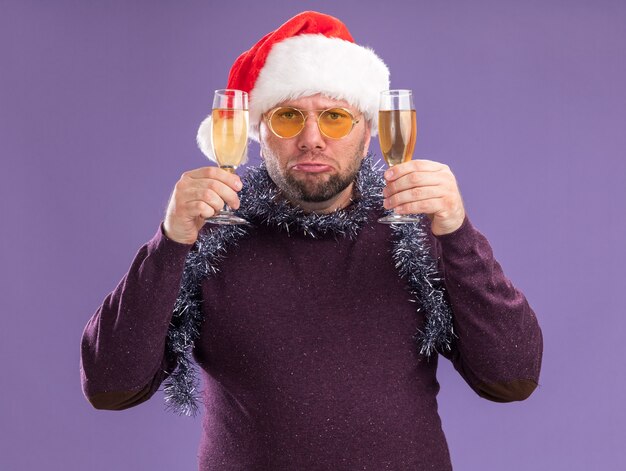 Triste hombre de mediana edad con gorro de Papá Noel y guirnalda de oropel alrededor del cuello con gafas sosteniendo dos copas de champán aislado en la pared púrpura