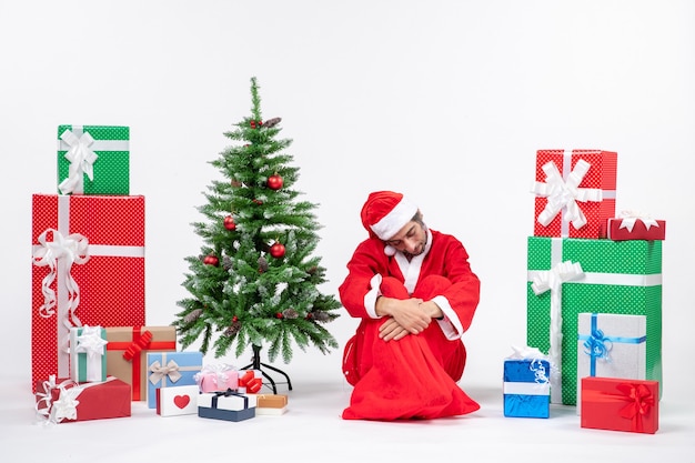 Triste infeliz joven vestido como Papá Noel con regalos y árbol de Navidad decorado sentado en el suelo sobre fondo blanco.