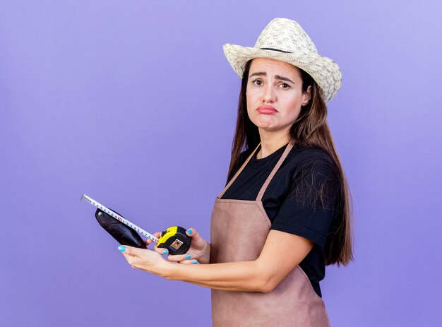 Triste jardinero hermosa chica en uniforme con sombrero de jardinería medir berenjena con cinta métrica aislado sobre fondo azul.