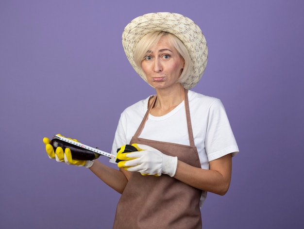 Triste jardinero rubio de mediana edad mujer en uniforme con sombrero y guantes de jardinería midiendo berenjena con medidor de cinta