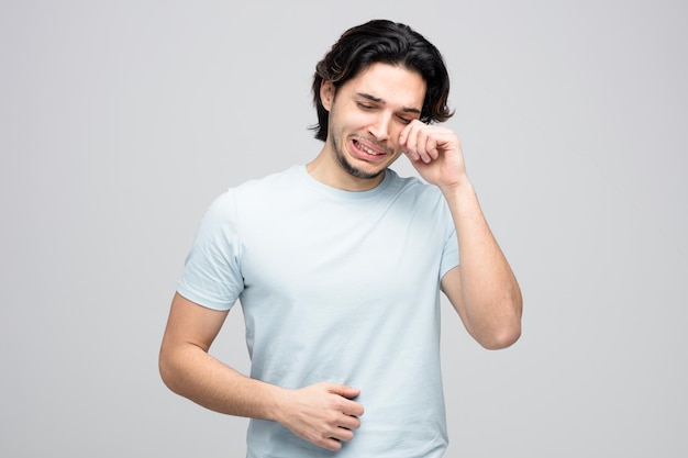 Triste joven apuesto llorando con los ojos cerrados y limpiándose el ojo con la mano aislado en el fondo blanco