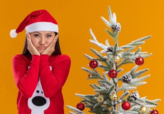 Triste joven asiática con sombrero de navidad con suéter de pie cerca del árbol de navidad poniendo las manos en las mejillas aisladas sobre fondo naranja