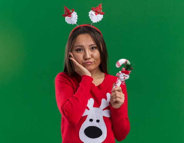 Triste joven asiática vistiendo un aro de pelo de Navidad con un suéter sosteniendo dulces de Navidad poniendo la mano en la mejilla aislado sobre fondo verde