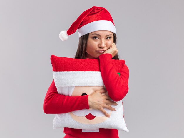 Triste joven asiática vistiendo gorro de navidad con suéter sosteniendo la almohada de navidad poniendo la mano en la mejilla aislado sobre fondo blanco.