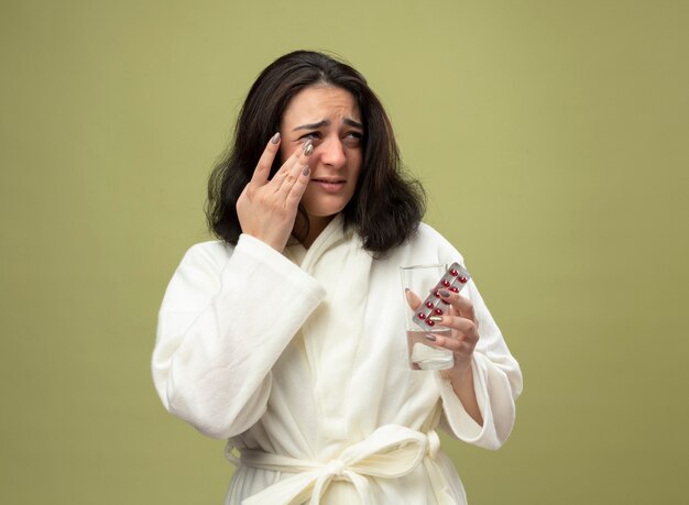 Triste joven caucásica enferma vestida con bata sosteniendo un vaso de agua y un paquete de píldoras médicas mirando de lado limpiando la oreja aislada sobre fondo verde oliva con espacio de copia