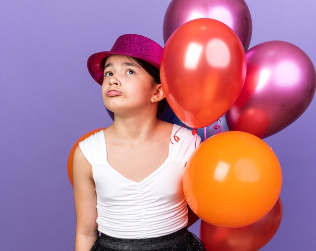 Triste joven caucásica con gorro de fiesta violeta sosteniendo globos de helio y mirando hacia arriba aislado en la pared púrpura con espacio de copia