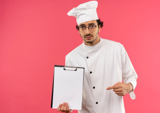 Triste joven cocinero vistiendo uniforme de chef y gafas sosteniendo y apunta a clipbord