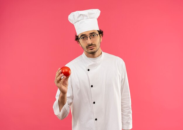 Triste joven cocinero vistiendo uniforme de chef y gafas sosteniendo tomate