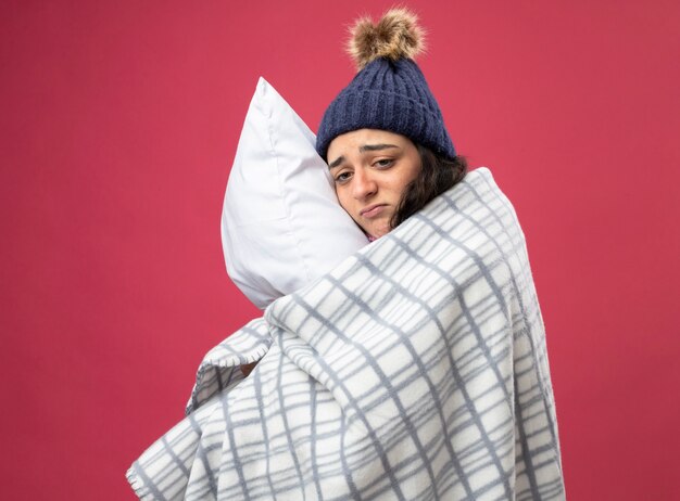 Triste joven enferma con bata de invierno sombrero y bufanda envuelta en cuadros de pie en la vista de perfil abrazando la almohada mirando al lado aislado en la pared rosa