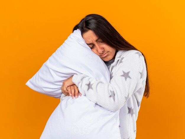 Triste joven enferma mirando hacia abajo abrazando la almohada aislada sobre fondo amarillo