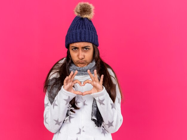 Triste joven enferma con sombrero de invierno con bufanda mostrando gesto de corazón aislado en rosa